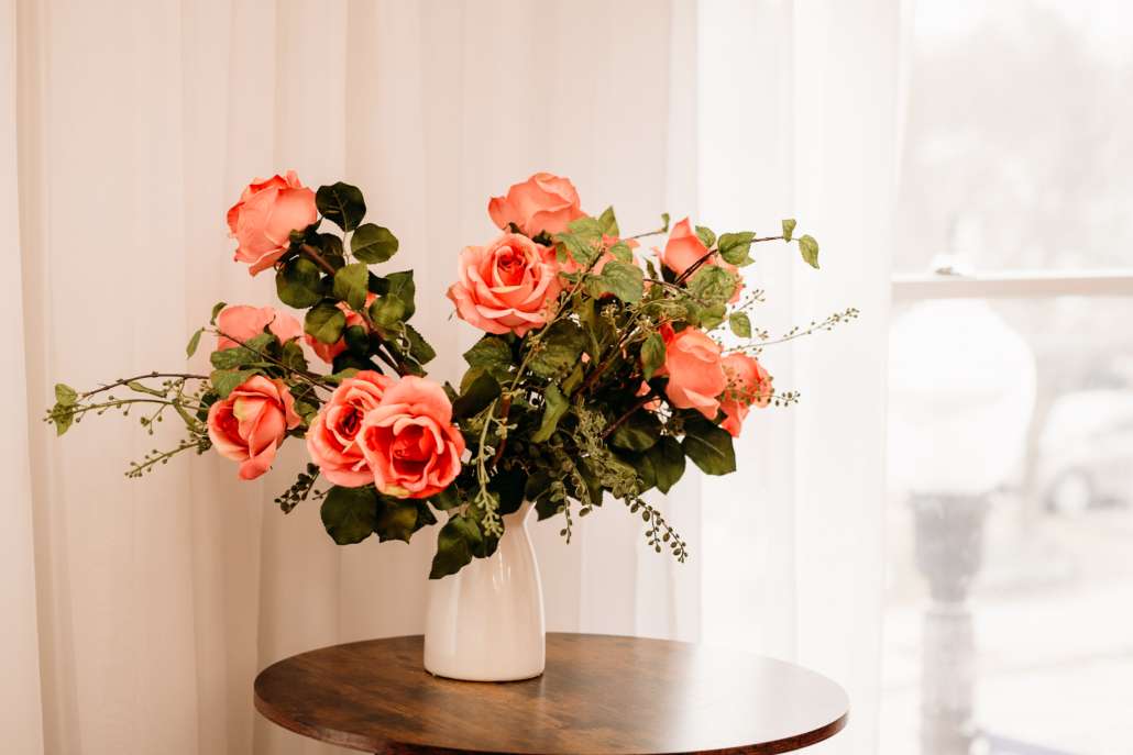 Close-up of a white ceramic vase with a lush arrangement of coral and peach roses on a dark wood pedestal table in a sunlit photography studio.
