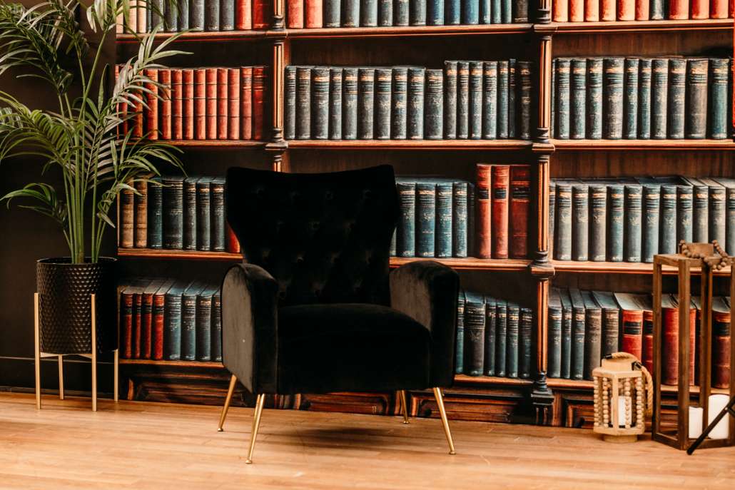 Dark academic photography set featuring floor-to-ceiling vintage books, a black velvet armchair, and warm wood flooring.