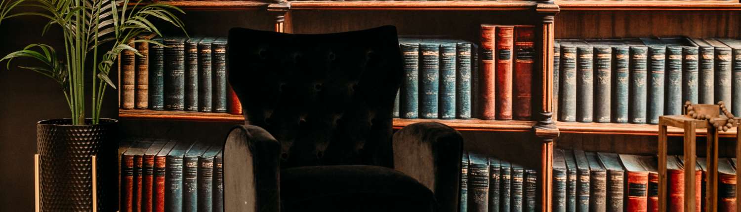 Dark academic photography set featuring floor-to-ceiling vintage books, a black velvet armchair, and warm wood flooring.