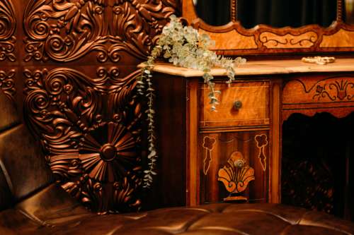 Close-up of a vintage dark wood vanity with intricate carvings and a brass drawer pull, paired with a tufted brown leather bench in a moody, Victorian-style photography studio.
