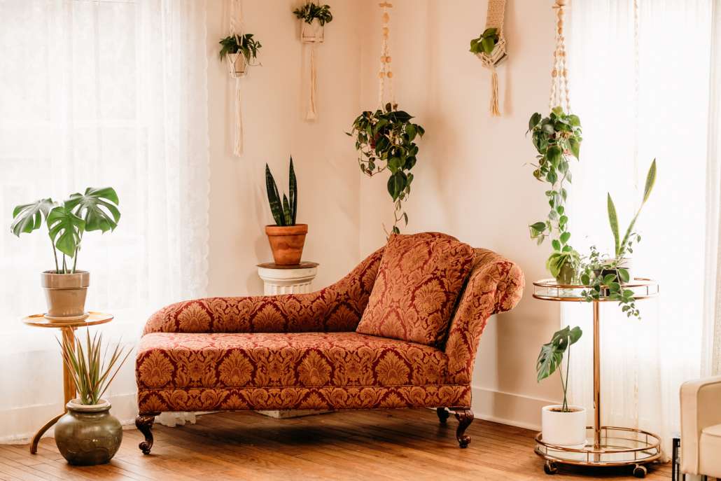 Vintage-inspired photography set featuring a red patterned chaise lounge, a gold tiered bar cart with potted plants, and macrame wall hangings in a bright, sunlit studio corner.