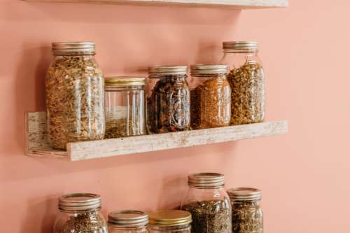Apothecary-style photography prop set featuring glass mason jars filled with dried botanicals and herbs on rustic wooden floating shelves against a soft peach wall.