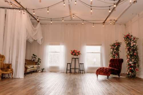 Spacious natural light photography studio at MI Studio Space featuring tall floral arches, a rust velvet chaise lounge, and overhead globe string lights on rustic wood floors.