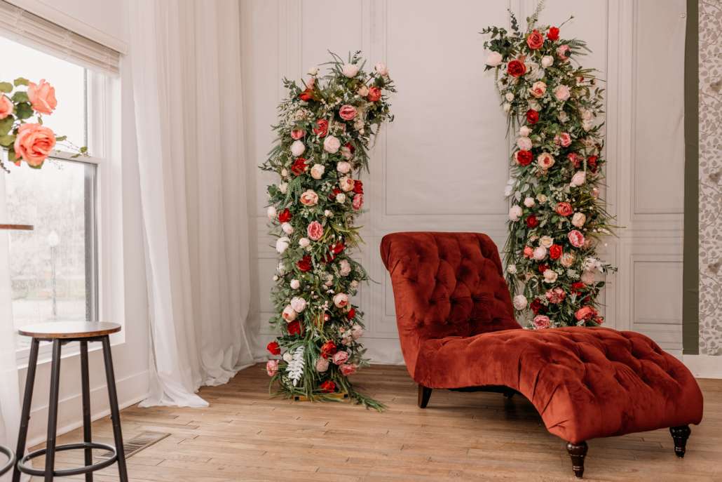 Elegantly styled photography set featuring a rust-colored velvet chaise lounge centered between two tall floral pillars with red and white roses at MI Studio Space.