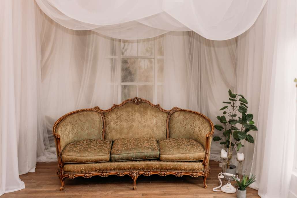 Ornate gold Victorian-style sofa centered in front of a floor-to-ceiling white fabric backdrop with sheer white curtains, creating a soft, ethereal photography set.