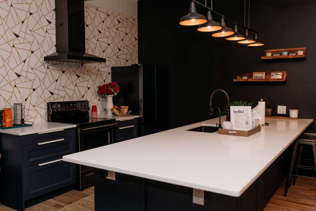Spacious lifestyle photography studio featuring a modern kitchen with navy cabinets, a white fireplace mantel with floral decor, and a cozy seating area with a white sofa and patterned rug.