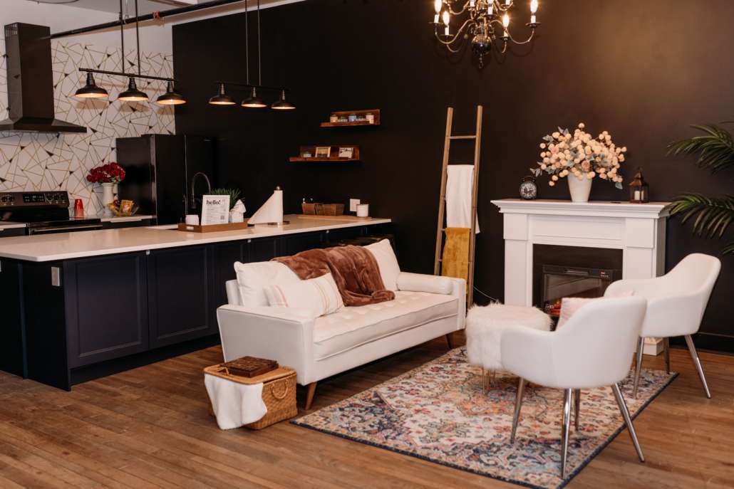 Spacious lifestyle photography studio featuring a modern kitchen with navy cabinets, a white fireplace mantel with floral decor, and a cozy seating area with a white sofa and patterned rug.