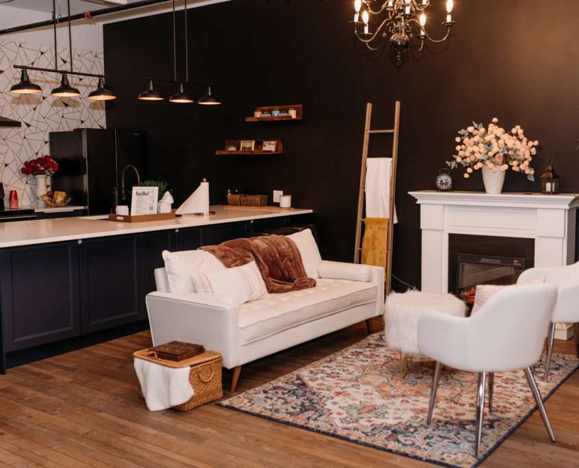 Spacious lifestyle photography studio featuring a modern kitchen with navy cabinets, a white fireplace mantel with floral decor, and a cozy seating area with a white sofa and patterned rug.