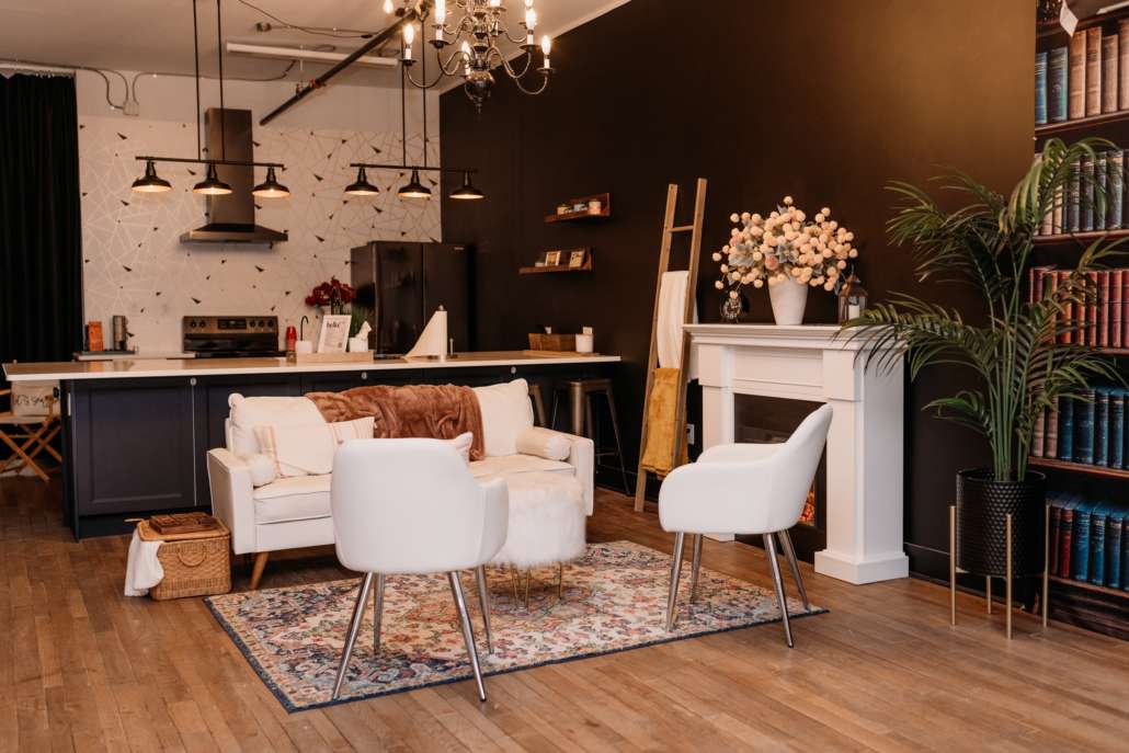 Spacious lifestyle photography studio featuring a modern kitchen with navy cabinets, a white fireplace mantel with floral decor, and a cozy seating area with a white sofa and patterned rug.