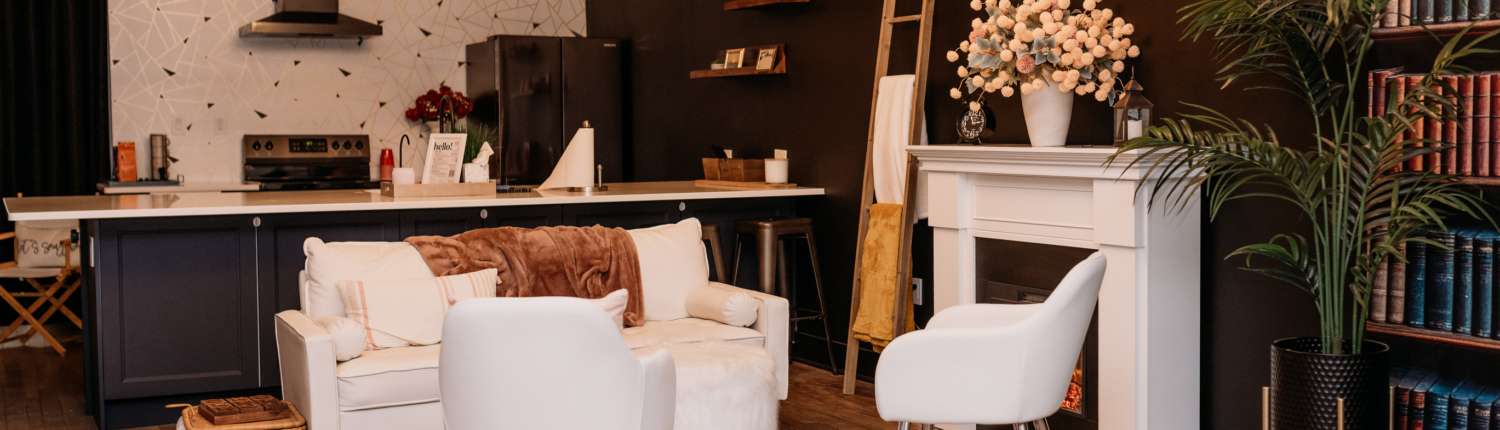 Spacious lifestyle photography studio featuring a modern kitchen with navy cabinets, a white fireplace mantel with floral decor, and a cozy seating area with a white sofa and patterned rug.