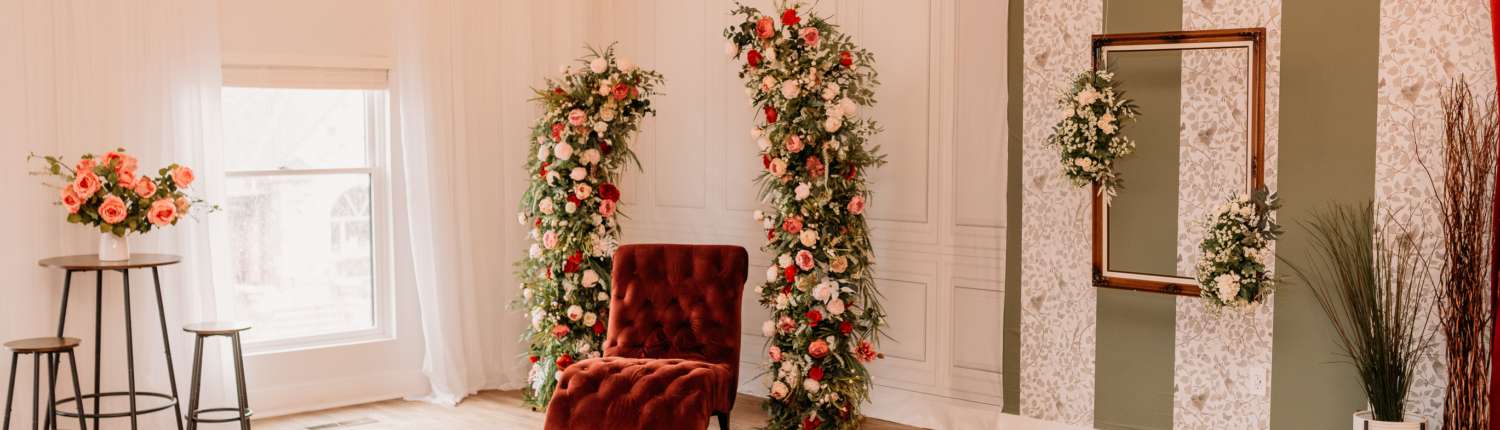Spacious natural light photography studio at MI Studio Space featuring two tall floral arches, a red velvet armchair, and a bistro seating area with overhead globe string lights on wood floors