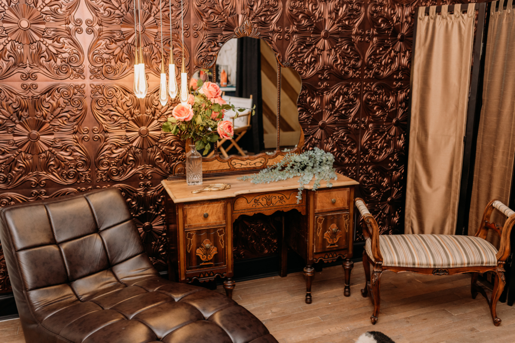 Moody Victorian-style photography set with a vintage wood vanity, a triptych mirror, and a black leather chaise lounge against a decorative embossed copper-patterned wall. Suggested Alt Text "Moody Victorian-style photography set with a vintage wood vanity, a triptych mirror, and a black leather chaise lounge against a decorative embossed copper-patterned wall.