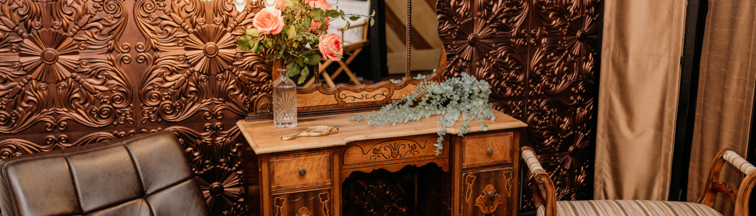Moody Victorian-style photography set with a vintage wood vanity, a triptych mirror, and a black leather chaise lounge against a decorative embossed copper-patterned wall. Suggested Alt Text "Moody Victorian-style photography set with a vintage wood vanity, a triptych mirror, and a black leather chaise lounge against a decorative embossed copper-patterned wall.