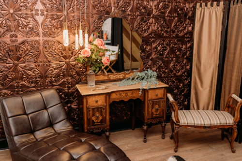 Moody Victorian-style photography set with a vintage wood vanity, a triptych mirror, and a black leather chaise lounge against a decorative embossed copper-patterned wall. Suggested Alt Text "Moody Victorian-style photography set with a vintage wood vanity, a triptych mirror, and a black leather chaise lounge against a decorative embossed copper-patterned wall.