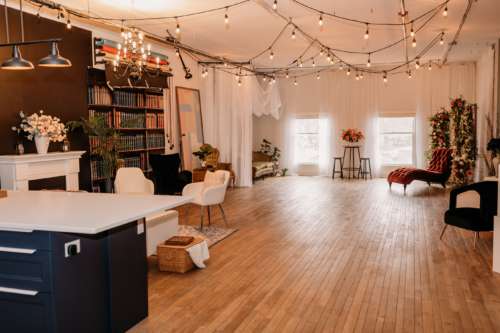 Spacious lifestyle photography studio featuring a modern kitchen with navy cabinets, a white fireplace mantel with floral decor, and a cozy seating area with a white sofa and patterned rug.