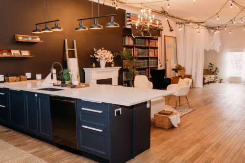 Spacious lifestyle photography studio featuring a modern kitchen with navy cabinets, a white fireplace mantel with floral decor, and a cozy seating area with a white sofa and patterned rug.