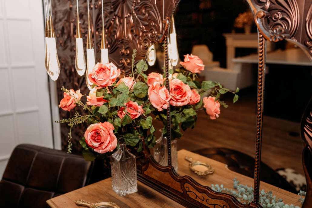 Moody Victorian-style photography set with a vintage wood vanity, a triptych mirror, and a black leather chaise lounge against a decorative embossed copper-patterned wall. Suggested Alt Text "Moody Victorian-style photography set with a vintage wood vanity, a triptych mirror, and a black leather chaise lounge against a decorative embossed copper-patterned wall.