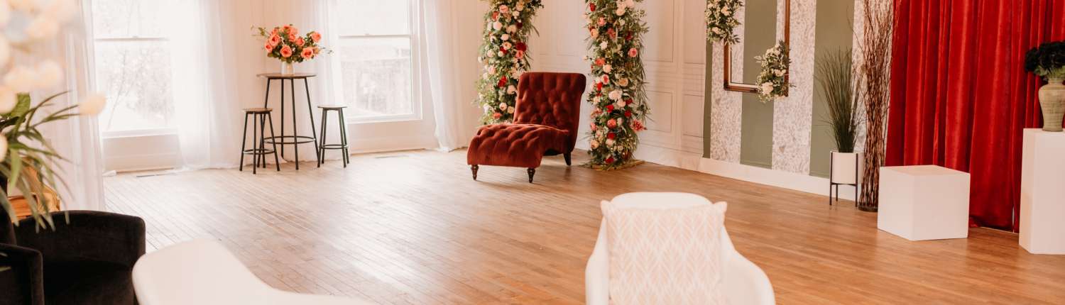 Spacious natural light photography studio at MI Studio Space featuring two tall floral arches, a red velvet armchair, and a bistro seating area with overhead globe string lights on wood floors