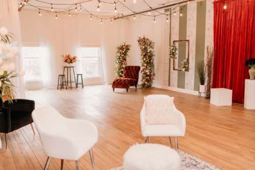 Spacious natural light photography studio at MI Studio Space featuring two tall floral arches, a red velvet armchair, and a bistro seating area with overhead globe string lights on wood floors