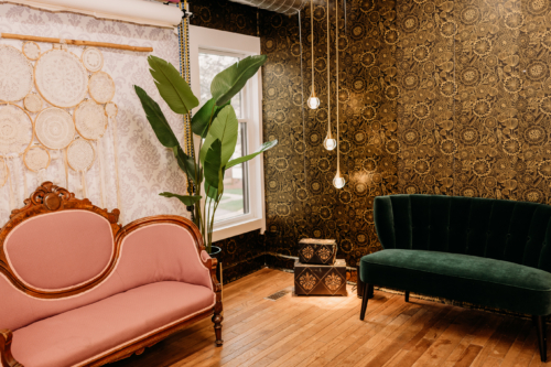 Boudoir photography set at MI Studio Space featuring a vintage pink velvet settee and a modern emerald green channel-tufted sofa against dark ornate wallpaper with hanging Edison bulb lights.