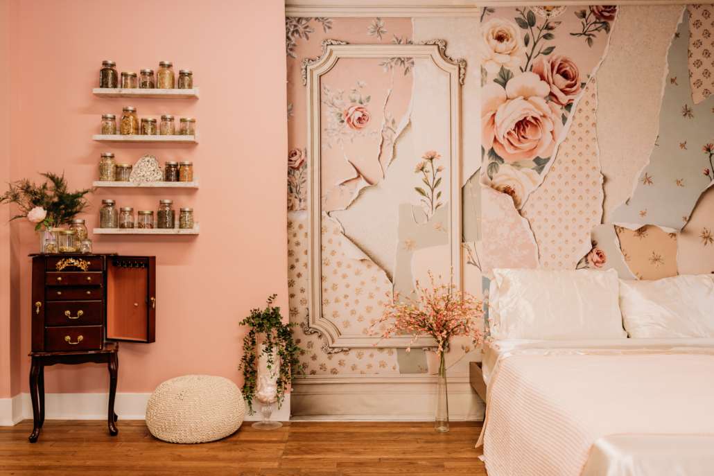Vintage-style boudoir photography set at MI Studio Space featuring a dark wood vanity with a triptych mirror, floating shelves with apothecary jars, and a pink floral wallpaper mural next to a white bed.