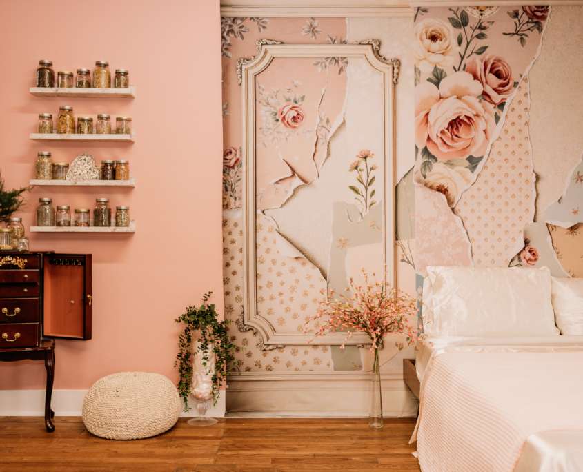 Vintage-style boudoir photography set at MI Studio Space featuring a dark wood vanity with a triptych mirror, floating shelves with apothecary jars, and a pink floral wallpaper mural next to a white bed.