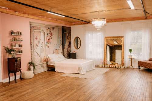 Spacious boudoir photography studio featuring a white paneled closet door, a minimalist bed set, vintage mirrors, and a crystal chandelier over polished wood floors.